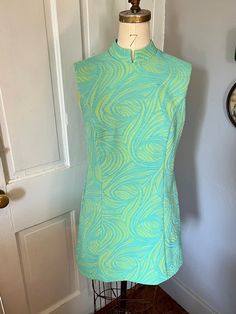 "This is a fantastic mini dress from the 1960s in the iconic mod silhouette, made by the brand Koret of California. Just imagine it with a pair of white go-go boots, or some colored leggings and platforms! The fabric seems to be a double-knit synthetic that has some stretch to it, with a raised aqua blue pattern on top of a sherbety lime green. There is a satin-like lining at the top near the collar and bust, but the rest of the dress is unlined. The dress is fitted at the waist then flares out, Coloured Leggings, 1960s Mod, Green Texture, Dress Tunic, Mini Shift Dress, Double Knitting, White Mini Dress, Mandarin Collar, Blue Pattern