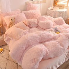 a bed with pink fluffy comforters and pillows on top of it, next to a teddy bear