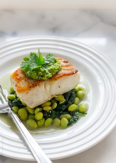 a white plate topped with fish and green peas next to a silver fork on top of a marble table