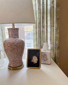 a pink vase sitting on top of a white table next to a lamp and photo