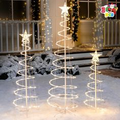 three lighted christmas trees in front of a house with snow on the ground and lights around them