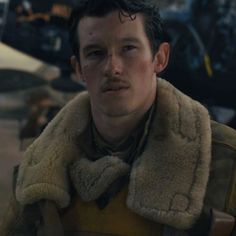 a young man in a fur collar and leather jacket looks at the camera while standing next to an airplane