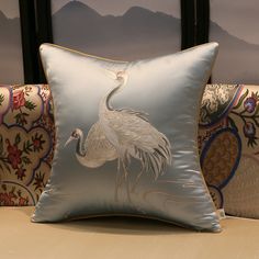 a blue pillow with a bird on it sitting next to a couch in front of a window