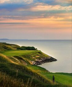 a person standing on top of a green golf course next to the ocean at sunset