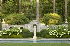 an outdoor fountain surrounded by bushes and flowers in the middle of a garden with trees