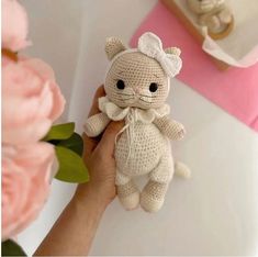 a hand holding a small crocheted teddy bear in front of pink flowers and other items