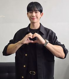 a young man making a heart shape with his hands while standing in front of a black couch