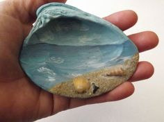 a hand is holding a small rock with shells on the beach and water in it