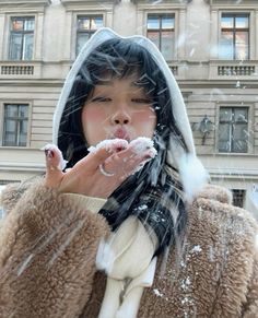 a woman standing in front of a building while blowing snow on her hands and covering her mouth