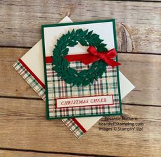 a christmas card with a green wreath on top and red ribbon around the edge, sitting on a wooden surface