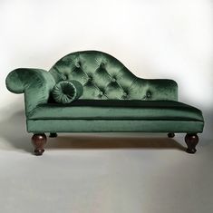 a green velvet chaise lounger with wooden legs and foot rests against a white wall