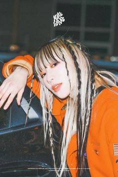 a woman with long hair and braids leaning on the hood of a car wearing an orange sweatshirt