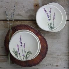 two white plates with purple flowers painted on them sitting on a wooden plate stand next to each other