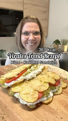 a woman with glasses is smiling in front of a large sandwich on a cutting board