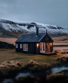 a small cabin in the middle of nowhere with snow covered mountains behind it and a stream running through the grass