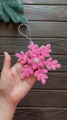 a hand holding a pink flower ornament on top of a wooden table next to a pine tree