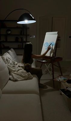 a woman sitting on a couch in front of an easel with a painting on it