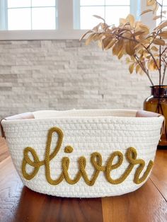 a white basket with the word silver written in gold thread on it sitting on a wooden table next to a potted plant