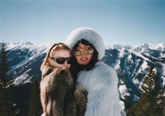 two women standing next to each other on top of a snow covered slope with mountains in the background