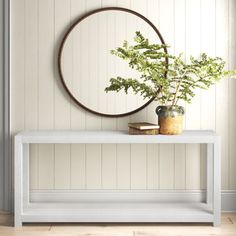 a white console table with a plant on top and a round mirror above it in a room