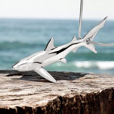 a silver shark necklace sitting on top of a piece of wood next to the ocean