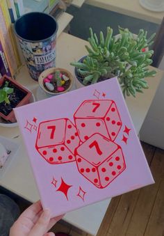 a person holding up a pink box with red dices on it and plants in the background