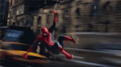 a spider - man leaps into the air in front of a car on a city street