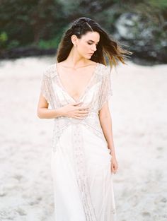 a woman standing on top of a sandy beach wearing a white dress and holding her hair in the wind