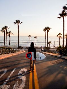 a person with a surfboard standing on the side of a road next to palm trees