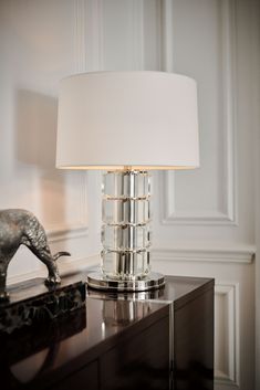 a table lamp on top of a wooden dresser next to a silver elephant statue and white shade