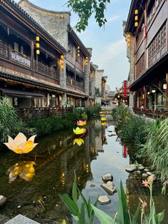 a river running through a lush green forest filled with lots of flowers next to tall buildings