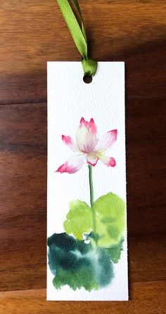 a watercolor painting of a pink and green flower on a white card hanging from a wooden table