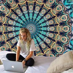 a woman sitting on a bed using a laptop computer in front of a colorful tapestry