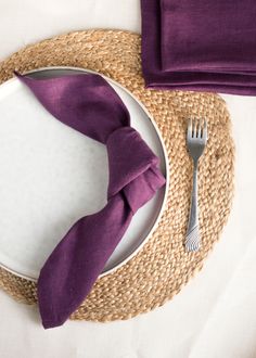 a place setting with purple napkins and silverware on a white table cloth covered plate