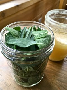 two jars filled with green leaves next to each other on a wooden table near a cup of orange juice