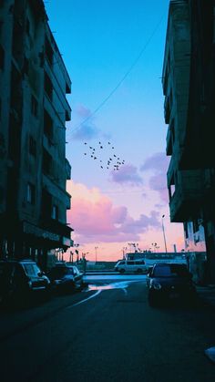 cars are parked on the side of an empty street at dusk with birds flying in the sky