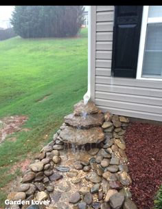 a small waterfall in the middle of a rock garden bed next to a house on a rainy day