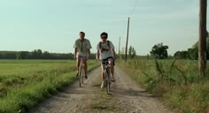 two people riding bikes down a dirt road in the middle of a green grass covered field