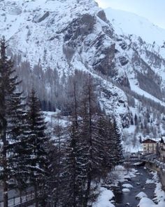 snow covered mountains and trees in the foreground with a river running through it, surrounded by buildings