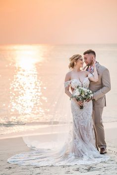 a bride and groom standing on the beach at sunset