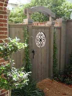 a wooden gate with an iron clock on it's face in the middle of a garden