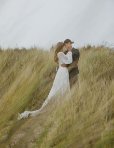 a bride and groom are standing in the tall grass on top of a hill with their arms around each other