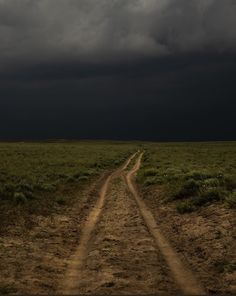 a dirt road in the middle of an open field under a dark sky with storm clouds