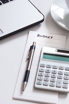 a desk with a calculator, pen and notebook on it next to a laptop