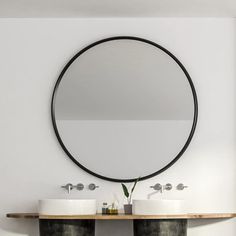 two white sinks sitting under a round mirror in a bathroom next to a wooden shelf