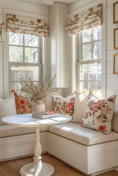 a corner bench with pillows and flowers on it in front of two windows that have white shutters