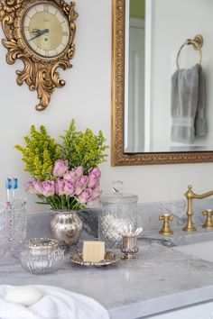 a bathroom counter with flowers, soap and other items sitting on it next to a mirror