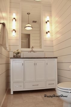 a white bathroom with two sinks and lights on the wall above it's mirror