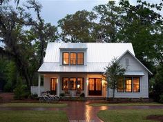 a white house with lots of windows and lights on it's front porch, surrounded by trees