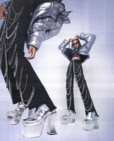 two women in silver and black outfits with chains on their legs, one wearing high heeled shoes